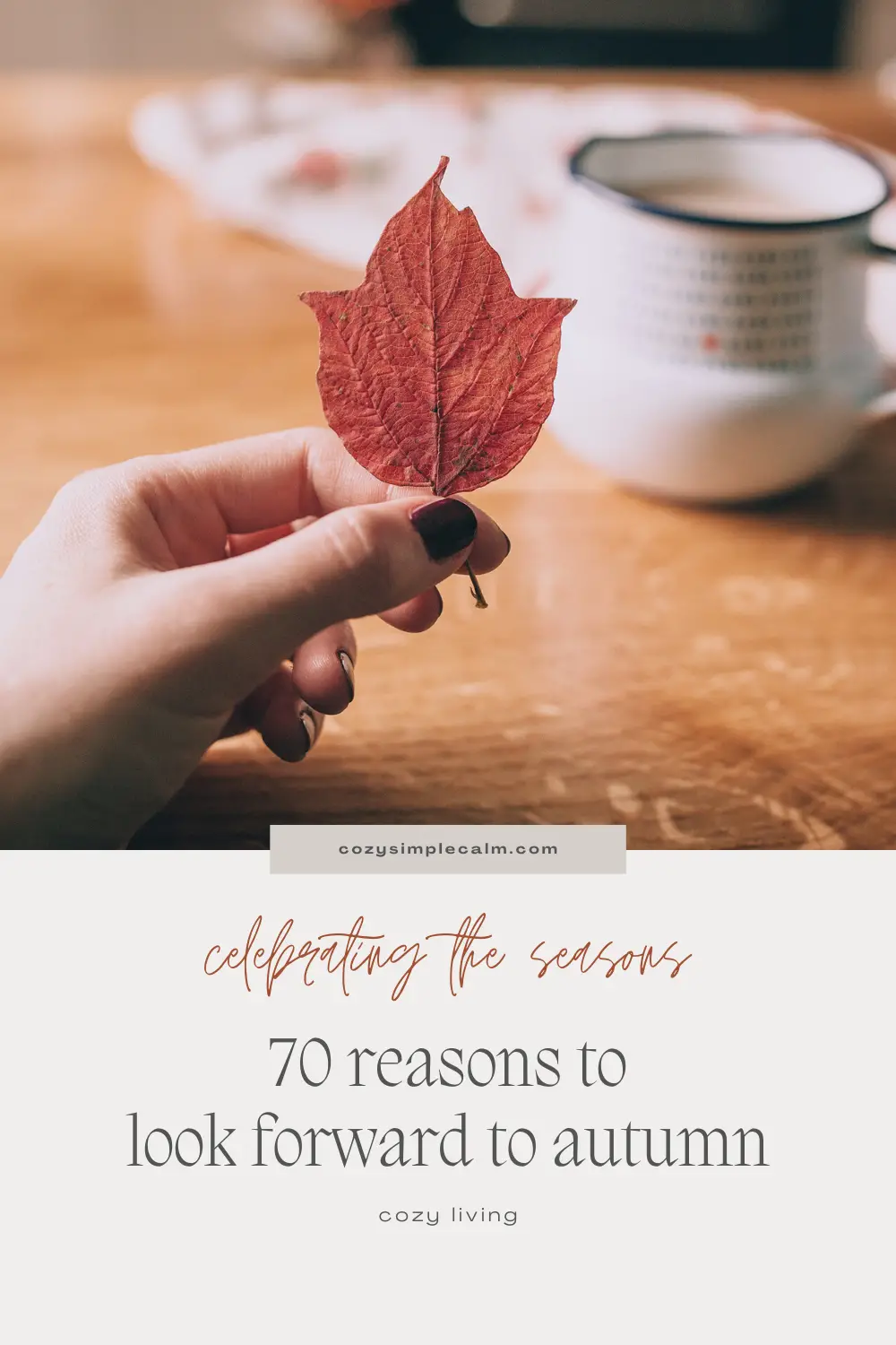 Image of hand holding red fall leaf in front of blurred coffee mug on table - Text overlay: Celebrating the seasons - 70 reasons to look forward to autumn - cozysimplecalm.com