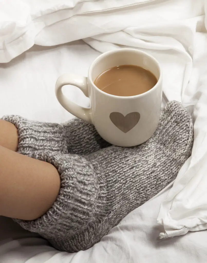 A cup of coffee or hot chocolate and female feet with socks on a white sheets.